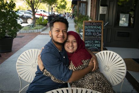 Two young adults sitting side by side hugging each other and smiling directly into the camera. The person on the left has short, dark hair and is wearing a denim button down shirt. The person on the right is wearing a patterned dress and a deep red head scarf. 