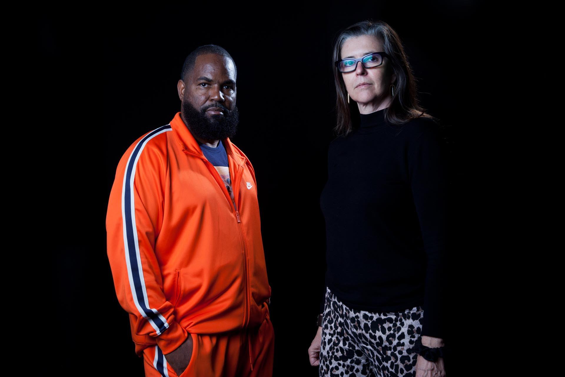 Black background with Black man wearing orange track suit and white woman wearing black sweater and glasses