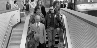 A black and white photograph of Stannous Flouride, wearing sunglasses and a leather jacket and boots, walking down the escalator at Montgomery BART station, surrounded by business people wearing suits.