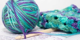 A ball of purple and turquoise yarn, a crochet hook and a crocheted square sitting on a flat surface