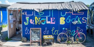 A photograph small structure with a corrugated metal roof, painted bright blue, with the words &quot;Jungle Books&quot; emblazoned colorfully on the side in Arabic, English and French. In front of the structure are a bike, a generator, and a chalkboard sign.