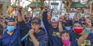 A group of people, many wearing surgical masks, stand outside in Chinatown and raise their fists as one person speaks into a microphone.