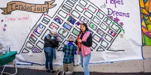 Three young people painting a mural on the street. One of the artists is holding a paintbrush while looking directly at the camera. The mural is a map of the Tenderloin neighborhood. 