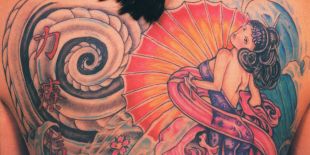 Close up photo of a full back tattoo on the upper part of a woman&#039;s back. The person has black hair that hits just below the shoulders to reveal a colorful display of Japanese symbols, characters and a female figure on the right side of the back. 
