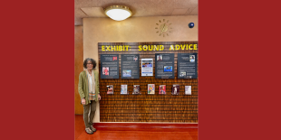 Color photograph of a woman standing to the left of a series of panels featuring images and text. Above the panels is a yellow title reading Exhibit: Sound Advice. There is a large, bright light hanging from the ceiling. 