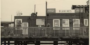 Sepia toned black and white photograph of a single story building sitting on a small pier. There are windows dotting the building and a fence in front of it. The white sign with greyscale lettering reads, Blanche's Place.
