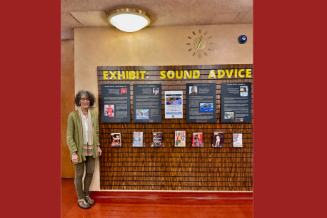 Color photograph of a woman standing to the left of a series of panels featuring images and text. Above the panels is a yellow title reading Exhibit: Sound Advice. There is a large, bright light hanging from the ceiling. 