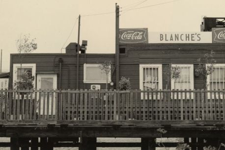 Sepia toned black and white photograph of a single story building sitting on a small pier. There are windows dotting the building and a fence in front of it. The white sign with greyscale lettering reads, Blanche's Place.