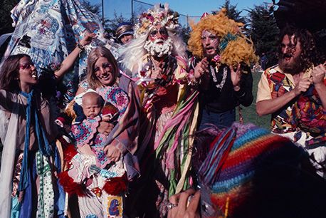 A lively group of people in colorful, elaborate costumes, wigs, and face paint pose together outdoors.
