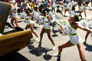 Girls with pom poms and matching sparkly outfits do a choreographed dance on a sunny paved street in Denver, Colorado.