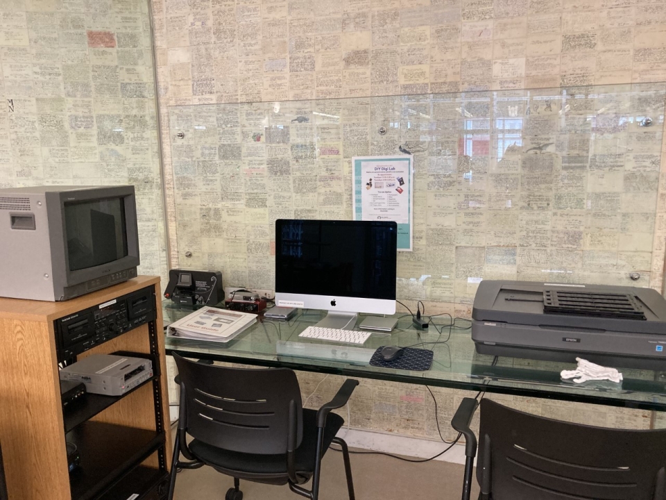 DIY Digi Lab room, including glasstop table with desktop computer, two chairs, flatbed scanner, cotton gloves, loose leaf binder, CRT monitor, and audiovisual playback equipment