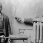 Black and white photograph from around 1906 shows African American inventor Charles S.L. Baker and another man, possibly Baker&#039;s brother Peter, standing behind heating (radiator) system. One man is holding a knob that is attached to two wires.
