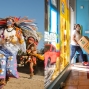 There are two images. On the left, participants in costume take part in the Dia de los Muertos festival in the Fruitvale District of Oakland. On the right, a person wearing a mask pushes four cardboard boxes on a dolly through the door.
