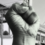 Black and white photo of a fist raised in the Black Power salute. In the background on the right a neon sign reading, &quot;American,&quot; above a marquee with the same name. On the left sits a white building with a tiled roof. 